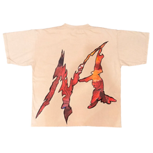 Load image into Gallery viewer, A cream-colored t-shirt with a large red and orange graphic print of the letter 'M' is laid flat on a gray carpet. The bottom of the frame shows a person's legs wearing gray slippers, black and white patterned shorts, and white socks, with a red bag at the bottom right.