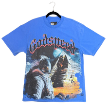 Load image into Gallery viewer, A blue short-sleeve graphic T-shirt laid flat on a carpeted floor, featuring a large space-themed print with the word 'Proudspy' in stylized lettering. A person's green-clad feet are visible at the top edge of the frame.