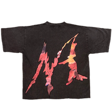 Load image into Gallery viewer, A black graphic t-shirt with a red, orange, and yellow abstract design, laid flat on a textured gray carpet. A person's legs wearing gray and black patterned pants and fluffy gray slippers are visible on the right side.