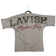 Load image into Gallery viewer, A white short-sleeve t-shirt laid flat on a brown carpeted floor, featuring 'LAVISH' in large black block letters with a white outline and 'Members Only' in red script. A pair of green superhero-style shoes is visible at the right edge of the frame.