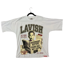 Load image into Gallery viewer, A white Lavish graphic t-shirt laid flat on a brown carpet, featuring a vintage-style newspaper collage print with a portrait and bold text. A person's feet in green gloves and white socks are visible on the right side of the frame.