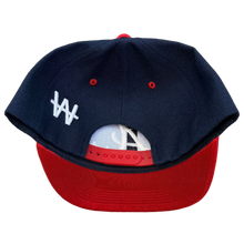 Load image into Gallery viewer, A navy blue and red snapback baseball cap with a white embroidered logo on the side, photographed from the back on a textured brown carpet background.