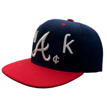 Load image into Gallery viewer, A navy blue and red baseball cap with white embroidered 'Atlanta' script and a small 'C' logo, resting on a textured carpet in a room with clothing racks in the background.