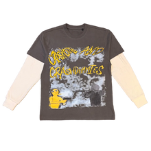 Load image into Gallery viewer, A dark gray long-sleeve t-shirt with a white long-sleeve layer underneath, featuring a graphic print that reads 'Crazy and Crash Dummies' in yellow over a distressed gray-and-white background with a yellow cartoon figure. The shirt is laid flat on a brown carpet with shoes visible at the top and bottom edges.
