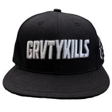Load image into Gallery viewer, A black flat-brim baseball cap with 'GRVTYKILLS' embroidered in white across the front, resting on a textured carpet in a room with clothing racks and a door in the background.
