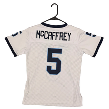 Load image into Gallery viewer, A white football jersey with 'MCCAFFREY' and the number '5' on the back, hanging on a black hanger over a brown carpet. A person's foot wearing a green cleat is visible at the bottom of the frame.

