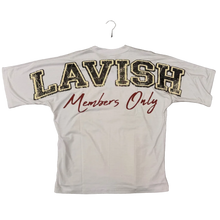 Load image into Gallery viewer, A white short-sleeve t-shirt laid flat on a brown carpeted floor, featuring 'LAVISH' in large gold and black outlined letters on the front and 'Members Only' in red script on the back. A person's green-clad feet are visible at the right edge.
