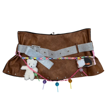 Load image into Gallery viewer, A brown suede-like belt bag or pouch laid flat on a textured gray carpet, decorated with a silver-and-white checkered strap, a colorful chain, a small white plush bear, and small accessories.
