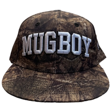 Load image into Gallery viewer, A camouflage baseball cap with 'MUGBOY' embroidered in white across the front, resting on a carpeted floor in a store.
