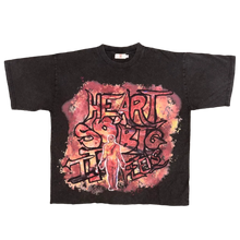 Load image into Gallery viewer, A black graphic t-shirt featuring the text 'HEART SHAPED INTO FIRE' in a stylized red, pink, and orange font, laid flat on a brown carpet. The photo is taken from a top-down perspective, with a person's feet in gray slippers visible at the bottom.

