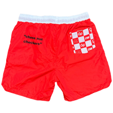 Load image into Gallery viewer, Red shorts with white checkered pattern and text on a gray surface, next to black Crocs shoes.
