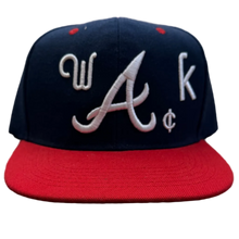 Load image into Gallery viewer, A navy blue and red baseball cap with white embroidered letters 'W', 'A', and 'k' on the front, resting on a textured carpet in a room with clothing racks in the background.
