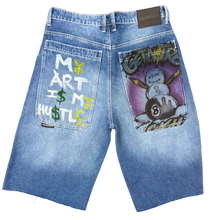 Load image into Gallery viewer, Blue jeans with a graphic design on the pocket laid flat on a carpeted floor.
